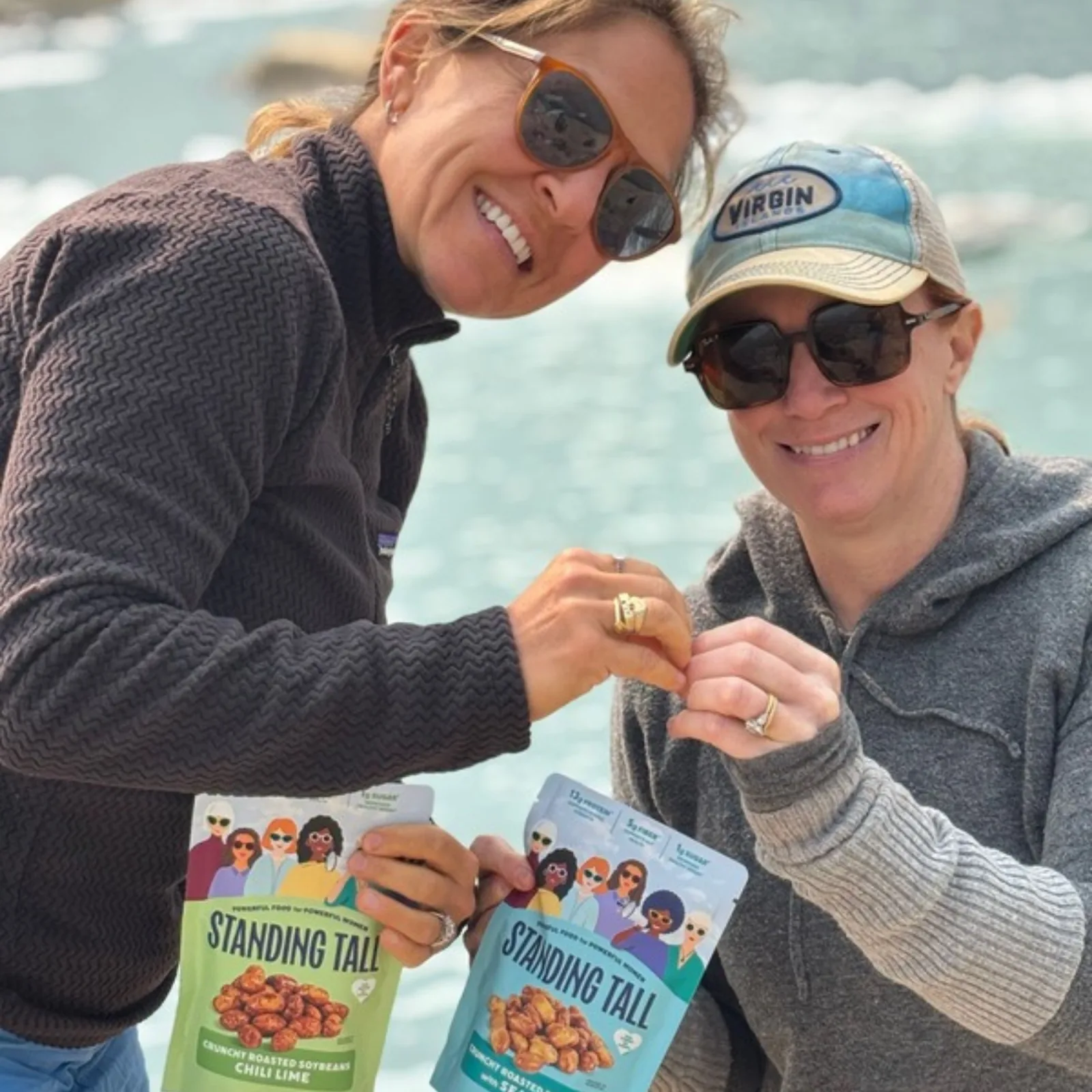 Two women outdoors by the water holding bags of Standing Tall Chili Lime and Sea Salt Crunchy Roasted Soybeans